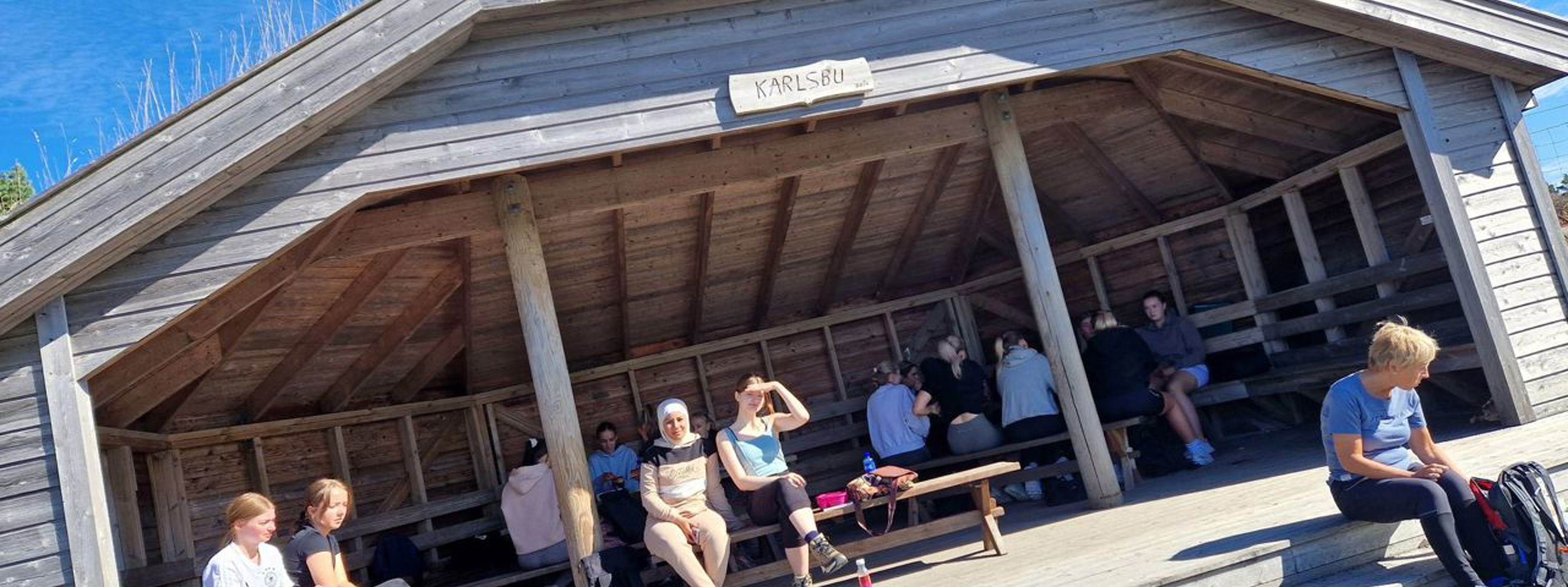 Elevar sit inne i ein trebygning. På skiltet øvst står det "Karlsbu". Det er sol og blå himmel.