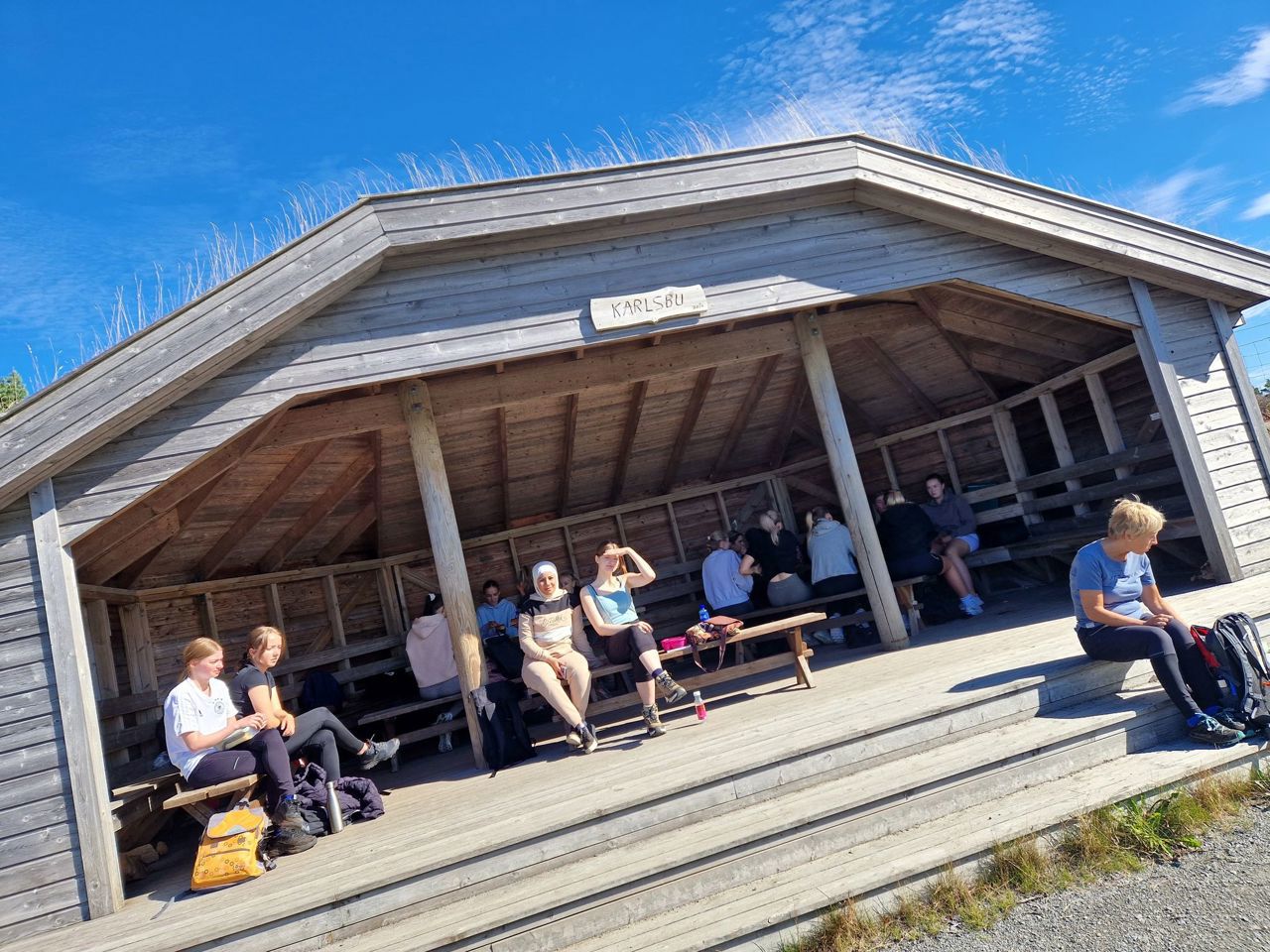 Elevar sit inne i ein trebygning. På skiltet øvst står det "Karlsbu". Det er sol og blå himmel.