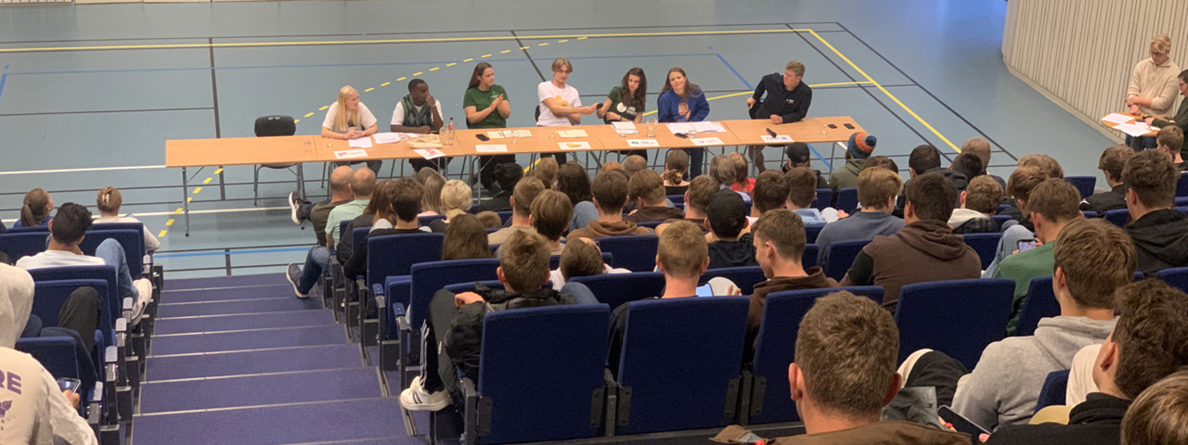 Politikarar diskuterer i Rubbhallen. Elevane høyrer på.