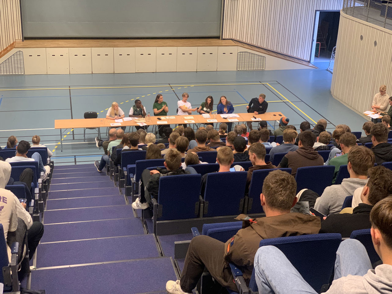 Politikarar diskuterer i Rubbhallen. Elevane høyrer på.