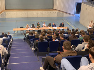 Politikarar diskuterer i Rubbhallen. Elevane høyrer på.